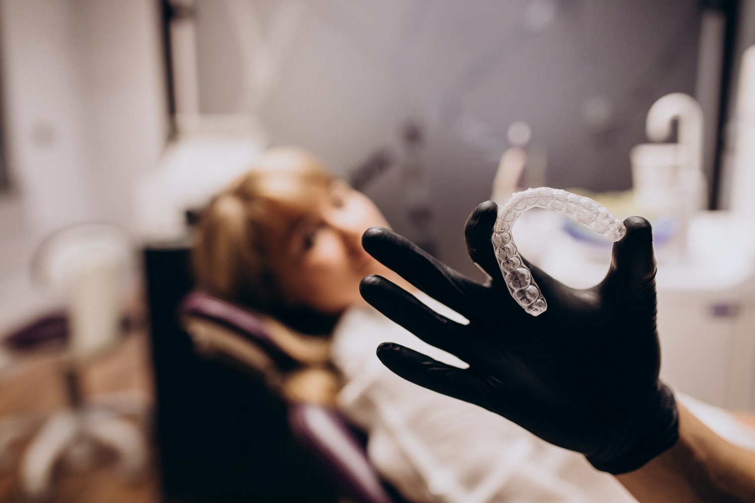 A gloved hand holding a clear dental aligner in the foreground, with a patient reclining in a dental chair softly out of focus in the background.